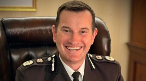 A smart-looking man with short brown hair and a black uniform with various metal symbols on the shoulders. He wears a white shirt and black tie underneath the main jacket. He is sitting on a brown leather chair and smiling at the camera.