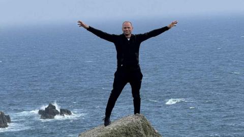 Tomas wearing a black jacket and black trousers- standing on a cliff with his arms spread out in the air - with the sea behind him