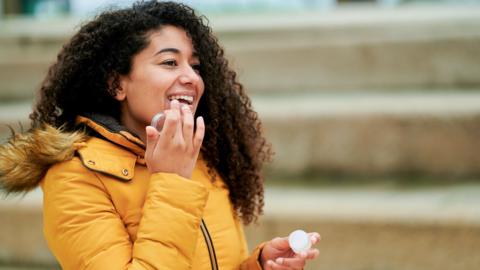 A young woman wearing a yellow puffer coat applies lip balm to her lips.