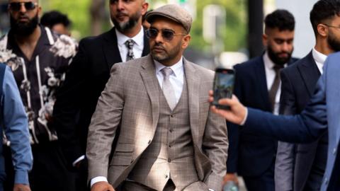 A man in a beige three-piece suit and a beige cap walks outside, surrounded by other men in suits. The man is wearing sunglasses and he has a neatly trimmed black beard.
