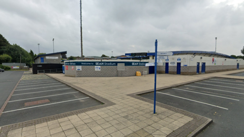 A view of a parking area and club stadium. No cars are parked there and the stadium is a white and grey building.