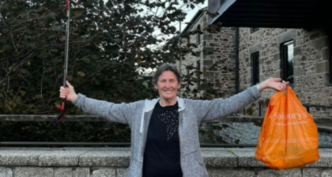 Image shows Ruth Major, who has short grey hair and is wearing a black jumper and grey cardigan, holding a litter grabber in her right hand and an orange Sainsbury's carrier bag in her left