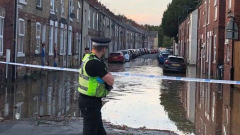 A policeman stands in front of a large pool of water which blocks a terraced street