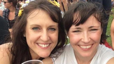 Two women, Chelsea Booth and Sharon Gorham, are outdoors on a grassy area, at an event. They are smiling, one wearing a flower crown, the other wearing a white top. There are various people gathered in the background.