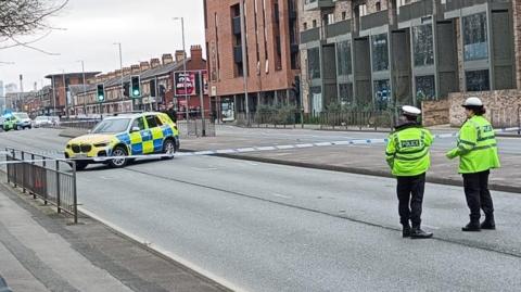 Two police officers in yellow uniform jackets and caps stand outside a corden across an emptied double carriageway. There are emergency services vehicles within the cordon.