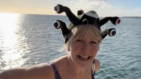 Ruth standing smiling by the sea wearing a swimming costume and a green and white knitted hat topped with 3d eyeballs. 