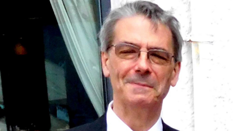 A headshot of Alan Hydes who is wearing tinted glasses, has short grey hair and is wearing a suit and tie, whilst smiling at the camera.