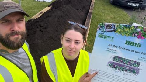 A photo of two people looking looking sad and pointing at a sign saying BBC Gardeners' World Live. They are both wearing high visibility jacket sand they are in front of a compost of soil. 