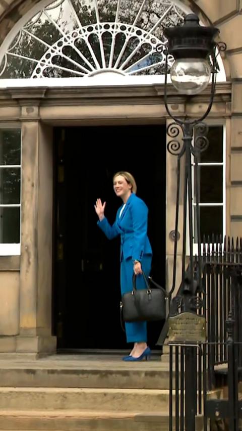 Jenny Gilruth is standing in the doorway of Bute House wearing a bright blue suit and waving.