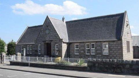 Insch Library - a grey brick building with large sloping roof behind a white fence, under a blue sky with some clouds.