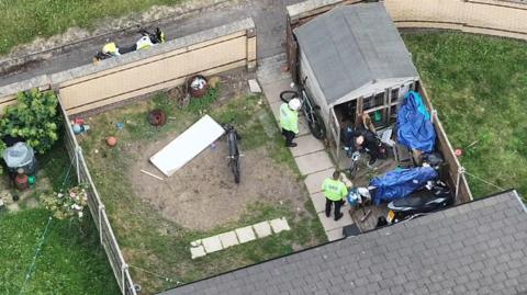 An aerial photo shows two police officers in uniform and another person who appears to be wheeling a bike out of a small outbuilding in a back garden. Two motorbikes and two bicycles can be seen. A police motorbike is parked up outside the garden.