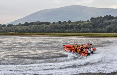 A boat goes speeding across the surface of the Solway with trees and hills in the background.