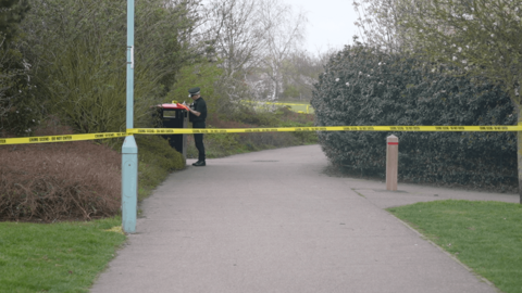 Yellow police tape attached to a lamppost. It is cordoning off an area of a park. A police officer is behind the tape and appears to be writing something down.