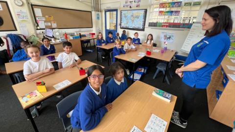 A primary school classroom full of children sitting at desks. Some of them are wearing bright blue school jumpers while some are wearing white t-shirts. There is a teacher stood at the front of the classroom wearing a blue polo shirt and smiling at the children with her hands clasped together.