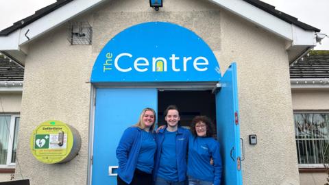 Rebecca Woods, Mitch Hill and Sharon Barnes stand in front of the entrance to The Centre. They are all wearing blue tops with The Centre branding. The door of the building is blue with the The Centre logo on the top.