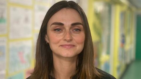 Charlene Hegarty has shoulder-length straight frown hair and small hoop gold earrings. Behind her out of focus a school corridor is visible.