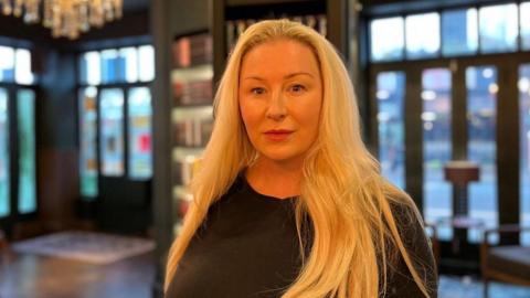 A woman with long blonde hair is wearing a black long sleeved top and is standing in front of shelves of books. 