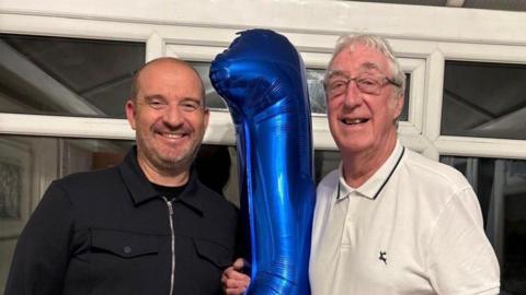 Two men one old and one younger smiling in a conservatory. They are holding a large helium birthday balloon.