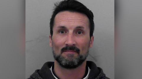 A mugshot of Mark Acklom. He has short dark hair and facial hair. He is pictured against a plain background with a blank expression. He is looking directly at the camera.