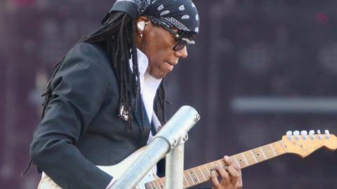 Nile Rodgers plays his guitar with ear phones in wearing suit and bandana
