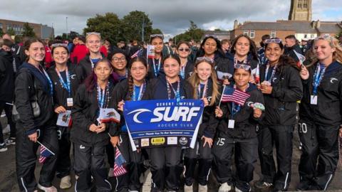 A girls’ team photo with a number of players wearing tracksuit tops and holding banners