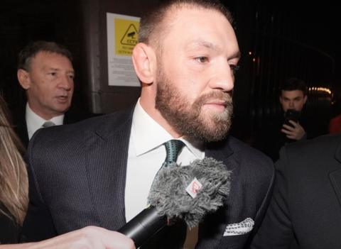 A man with a brown beard and short hair is facing away from a news microphone. He is wearing a dark suit, white shirt and dark green tie. 