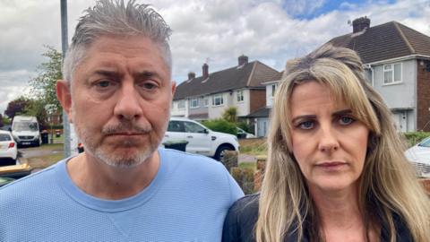 A sad looking couple, the man with grey hair and a blonde woman, are stood on a residential street.
