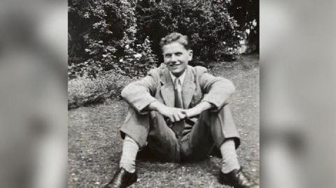 A black and white picture of Gordon wearing a light shirt, tie and smart trousers with a light coloured over blazer.  He is wearing black shoes and light socks and is smiling at the camera, with trees behind him. It was taken in his garden in 1951. 