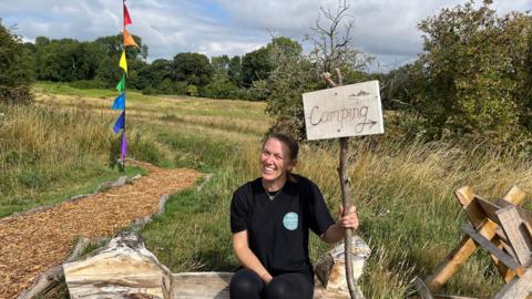A woman wearing all black looking at the camera and smiling. She is holding a sign made out of a plank of wood and a tree branch which reads "Camping". She is sitting on a homemade wooden bench made out of a large log. There is a field behind her and a path paved with wood chippings next to her.