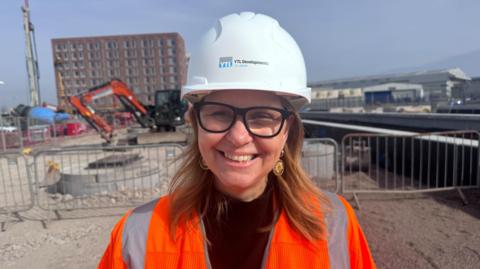 Helen Godwin is wearing a white hard hat and high vis jacket. She is pictured on a building site, smiling at the camera.
