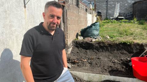 Tom Clarke, who has dark hair with silver speckles and a dark beard, smiles at the camera. He wears a black polo shirt and white shorts and his dug up garden can be seen in the background. 