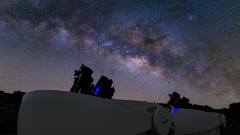 A night sky over several telescopes, black cylinders pointing towards the sky. The sky is brightly lit with stars and ranges in colours from deep blue to white.
