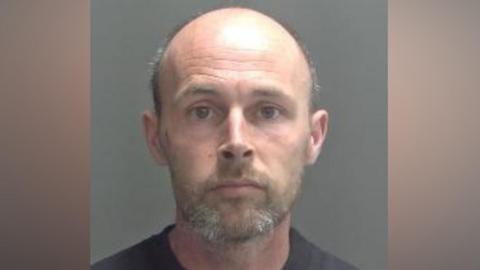 A police custody photo of Hudson. He has a short brown-and-grey beard and moustache and is bald. He is wearing a black T-shirt and looking into the camera.