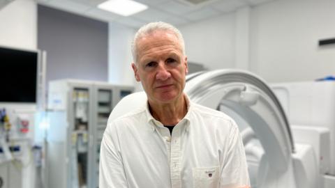 Tom Binet, a man with short white hair, is wearing a white shirt and no tie. He is standing in front of medical equipment, in a room which appears to be in a hospital. He is staring directly at the camera.