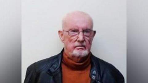 Cameron has short white receding hair and a goatee beard. He is wearing glasses and a black leather jacket.