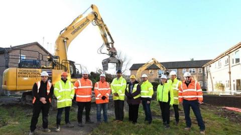 A group of people wearing hi-vis jackets and hard hats stand in a line on a greeb patch of land in front of a large yellow digger. Terraced houses can be seen in the background