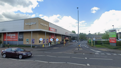 Wide view of Burghmuir Retail Park in Stirling showing Jollyes pet store, Waitrose and TK Maxx signs beside a car park and roadside junction under a partly cloudy sky.