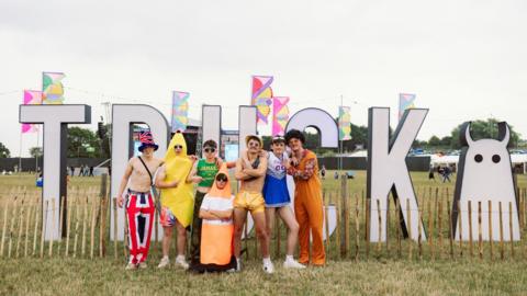 A group of seven young men stand in front of a large letter display in a field. The letters read 'Truck' and there is the Truck Festival logo and multicoloured festival flags in the backgrounds. From left to right the men are wearing various costumes. The first man is topless with a Union Jack top hat and matching trousers, with a stachel bag strap across his torso, next to him a man stands wearing a banana costume, the next man wears a green tank top with yellow writing which says 'Jamaica', another man is crouched on the ground which his arms crossed dressed like an orange and white traffic cone. Then standing is a man who is topless wearing golden shorts and a gold bucket hat, a man wearing a blue and white cheerleader's costume and a man in a 70s style outfit with a black afro wig.
