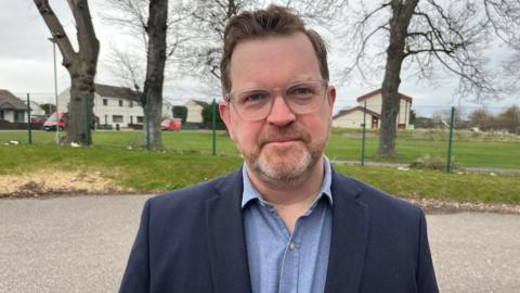 Picture of Calum MacPherson with his head and shoulders in the frame. He has brown hair, a grey bear and glasses and is wearing a dark blue blazer with a lighter blue shirt underneath.