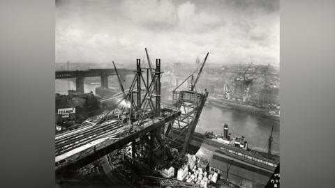 A black and white photograph showing the building on the Tyne Bridge from the Gateshead side. Construction is taking place on either side of the river, but the arch going over the water is just beginning to take shape. There are factories and buildings surrounding the structure, with the High Level Bridge and Swing Bridge visible behind it to the left. There are multiple cranes working on the bridge. The sky is covered in white clouds.