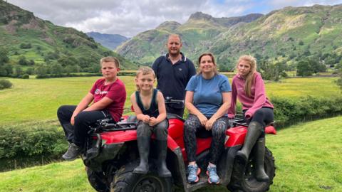 The Benson family sat on a red quad bike with the fells in the background. Dad Jonathan is standing behind the quad bike, while his two boys are on the left of the image, sat on the bonnet, mum Nicola is in the middle and their daughter is on the right, sat on the back. They are all in informal sportwear and wellies or trainers. They are looking at the camera and smiling.