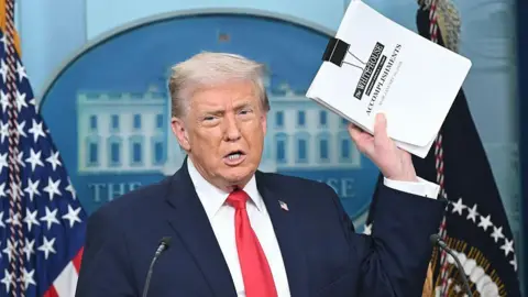 Donald Trump holds a document labeled 'White House Accomplishments' in the briefing room