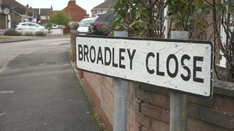 A picture of the road sign with houses in the back ground and parked cars.