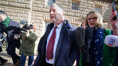 Mike Amesbury, wearing a navy blue coat over a white shirt and burgundy tie, walks away from court flanked by journalists holding microphones and camera operators