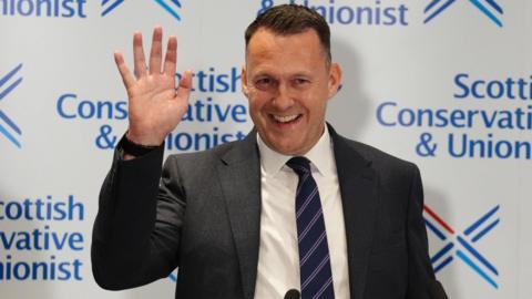 Russell FIndlay smiles and waves in front of a white backdrop with the party logo