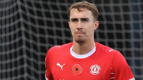 Lewis Billington in action for Crewe Alexandra. He is wearing a red home shirt and is jogging away from goal with the net visible, but blurred, in the background.