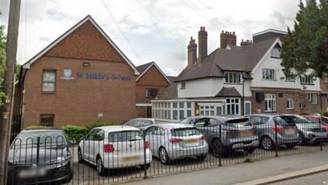 St Hilda's School seen from the outside with the school name in blue letters and a logo next to that. It looks like a large brick house rather than a school, standing next to a road and adjacent to another large white building with several chimneys that is presumably part of the school. There is a small car park with about 8 cars and a black fence.