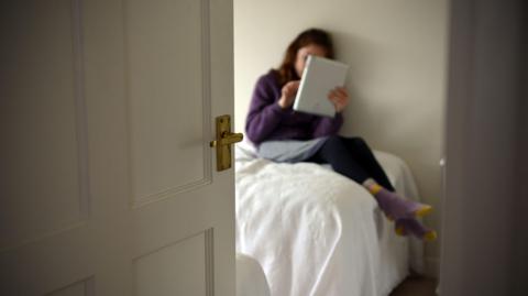 A young girl using a tablet computer on her bed, with the door open.
