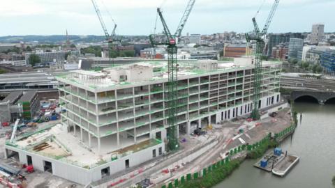 An aerial image of the new building under construction. The main frame of the building is complete, with three cranes beside the structure. It is a large rectangular building with five floors and is situated next to the river. The skyline landscape of the city can be seen in the background. 