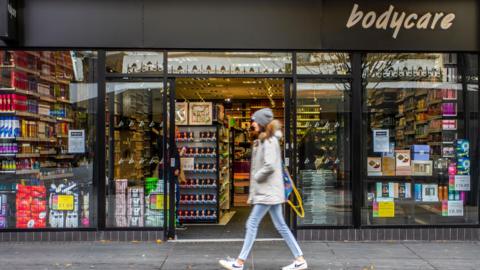 A person in a grey jacket and blue jeans walks past a shop with 'Bodycare' written on the top-right of the frontage. The store has large glass windows displaying various products such as toiletries, cosmetics, and household items. Shelves of products are visible inside the entrance. Signs on the windows advertise prices and promotions.